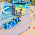 Blue and green satin Beach Please scrunchie phone wrist strap clipped to the matching Beach Please Wavy Luxe iPhone case with a beach chair and cocktail design, styled on a sandy shoreline background with starfish and ocean waves.