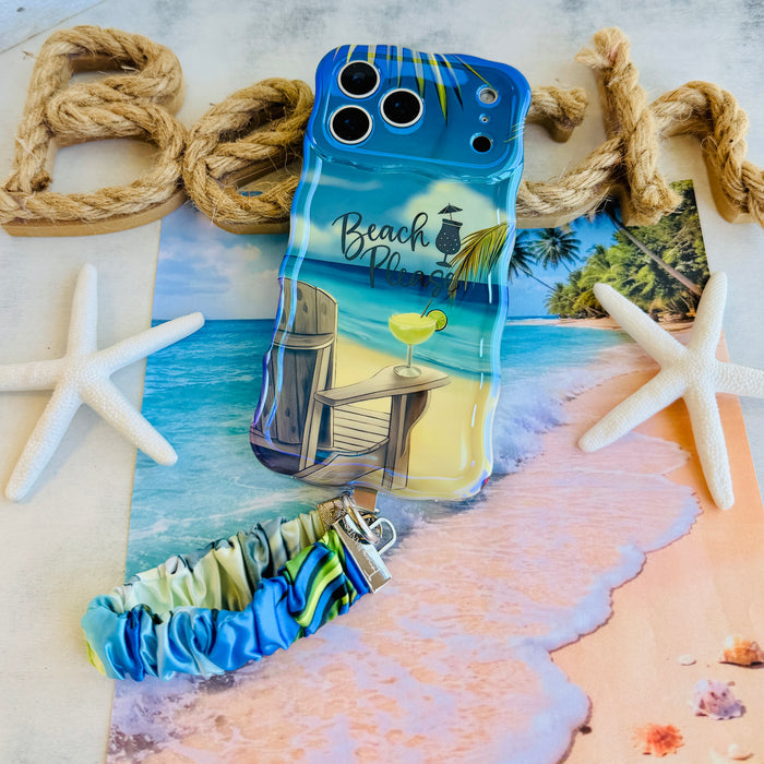 Blue and green satin Beach Please scrunchie phone wrist strap clipped to the matching Beach Please Wavy Luxe iPhone case with a beach chair and cocktail design, styled on a sandy shoreline background with starfish and ocean waves.