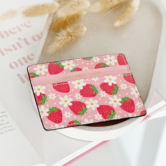 Pink card holder with strawberry and daisy prints, placed next to a book and dried flowers.