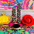 Bloom Bonita silicone Samsung Galaxy Watch band with Mexican folk art florals in coral, red, yellow, teal, and pink on a black base, shown displayed against a vibrant Mexican folk art backdrop with rose and dress accents.
