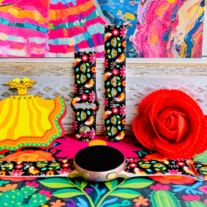 Bloom Bonita silicone Samsung Galaxy Watch band with Mexican folk art florals in coral, red, yellow, teal, and pink on a black base, shown displayed against a vibrant Mexican folk art backdrop with rose and dress accents.