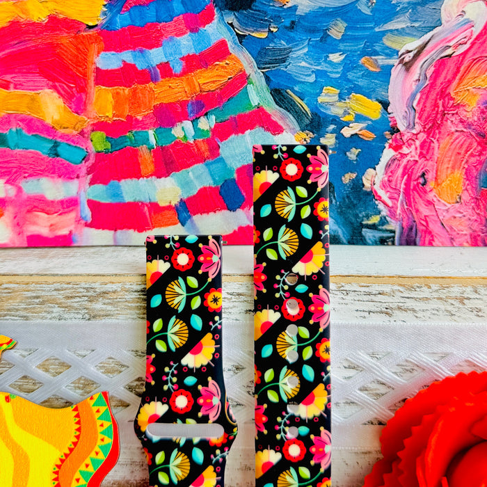 Bloom Bonita silicone Samsung Galaxy Watch band with Mexican folk art florals in coral, red, yellow, teal, and pink on a black base, shown displayed against a vibrant Mexican folk art backdrop with rose and dress accents.