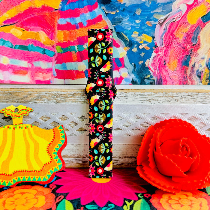 Bloom Bonita silicone Samsung Galaxy Watch band with Mexican folk art florals in coral, red, yellow, teal, and pink on a black base, shown displayed against a vibrant Mexican folk art backdrop with rose and dress accents.