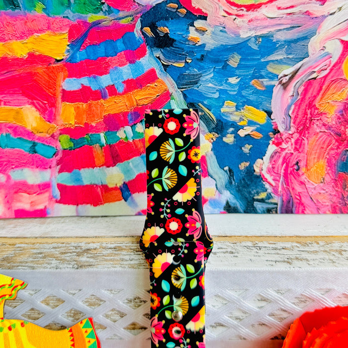 Bloom Bonita silicone Samsung Galaxy Watch band with Mexican folk art florals in coral, red, yellow, teal, and pink on a black base, shown displayed against a vibrant Mexican folk art backdrop with rose and dress accents.