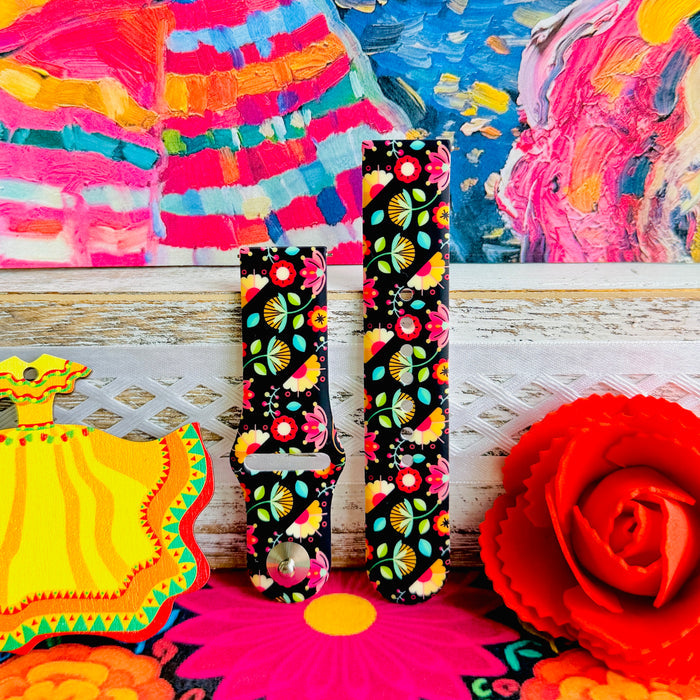 Bloom Bonita silicone Samsung Galaxy Watch band with Mexican folk art florals in coral, red, yellow, teal, and pink on a black base, shown displayed against a vibrant Mexican folk art backdrop with rose and dress accents.