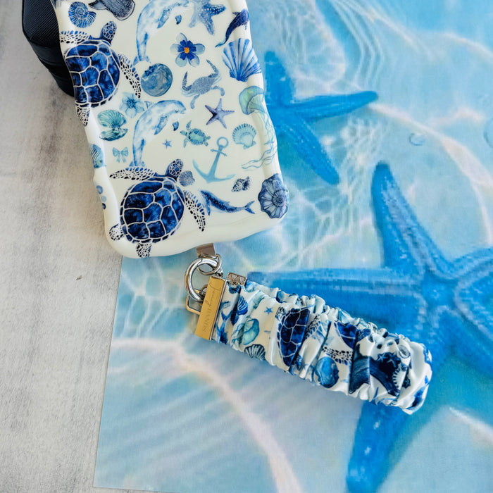 A Blue Lagoon scrunchie phone wrist strap with a blue and white sea turtle and ocean design attached to a matching Wavy Luxe case on a blue starfish background.