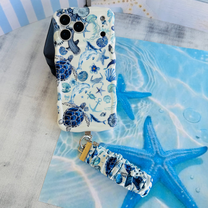 A Blue Lagoon scrunchie phone wrist strap with a blue and white sea turtle and ocean design attached to a matching Wavy Luxe case on a blue starfish background.