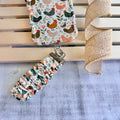 Cream and floral Cluckin' Cute scrunchie phone wrist strap with a hand-illustrated chicken and wildflower print, clipped to the matching farmhouse chicken Wavy Luxe iPhone case, styled on a rustic wood and burlap background.