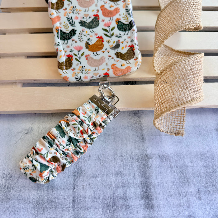 Cream and floral Cluckin' Cute scrunchie phone wrist strap with a hand-illustrated chicken and wildflower print, clipped to the matching farmhouse chicken Wavy Luxe iPhone case, styled on a rustic wood and burlap background.