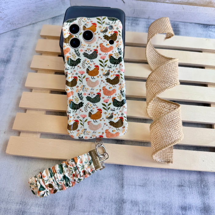 Cream and floral Cluckin' Cute scrunchie phone wrist strap with a hand-illustrated chicken and wildflower print, clipped to the matching farmhouse chicken Wavy Luxe iPhone case, styled on a rustic wood and burlap background.