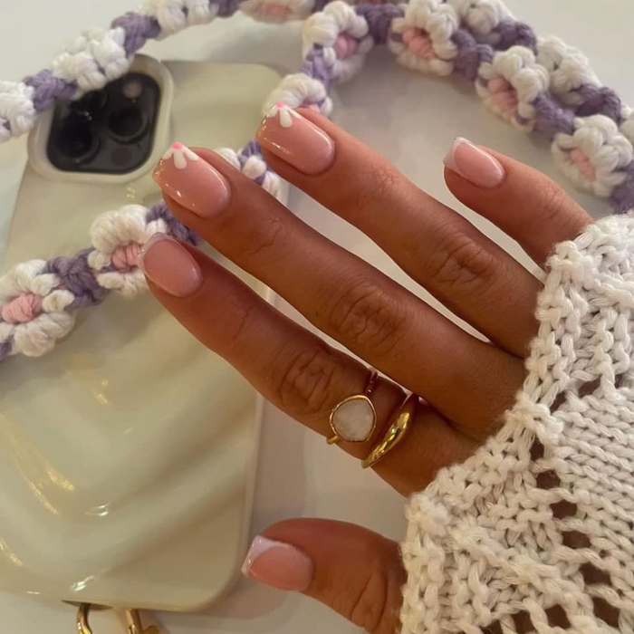 Hand with pastel pink floral nails, gold rings, and a lilac crochet flower phone lanyard.