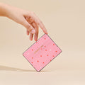Pink vegan leather card holder with red hearts pattern, held in a hand against a beige background.