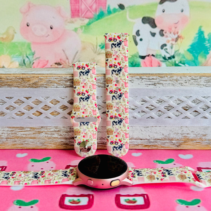 Farm Girl Cutie silicone Samsung Galaxy Watch band with dairy cows, red tractors, chickens, ducks, pink watering cans, strawberries, baskets, and roses on a cream base with pink gingham squares, shown displayed and worn against a farmhouse country backdrop.
