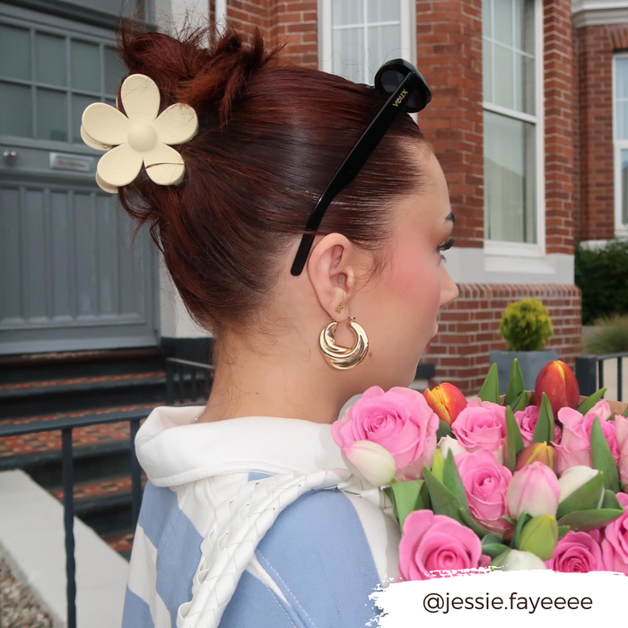 Woman wearing cream flower claw clip, gold hoops, and sunglasses holds pink flowers outside.