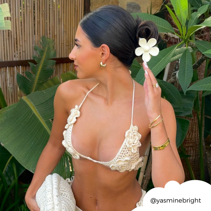 Woman in a crocheted bikini wears a cream flower-shaped claw clip, standing among green tropical plants.