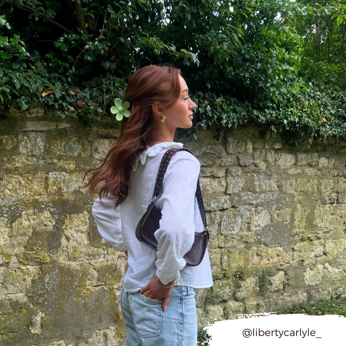 Woman with long hair wearing a green flower claw clip, white blouse, and jeans stands by a stone wall.
