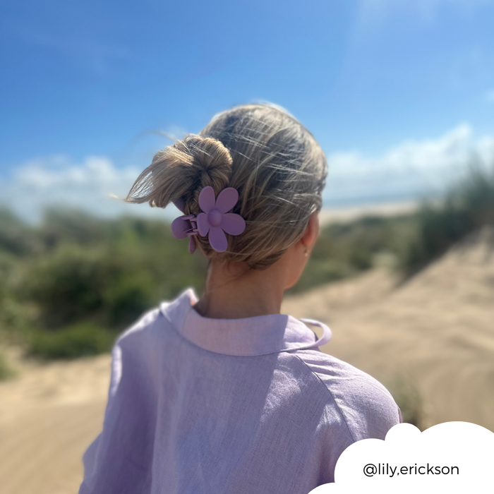 Person wearing a lavender shirt and lilac flower-shaped hair claw stands on sandy beach, facing grassy dunes.