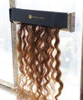 Wavy, brown hair extensions from Perfect Locks cascade from a sleek black box, illuminated by sunlight through a nearby window.