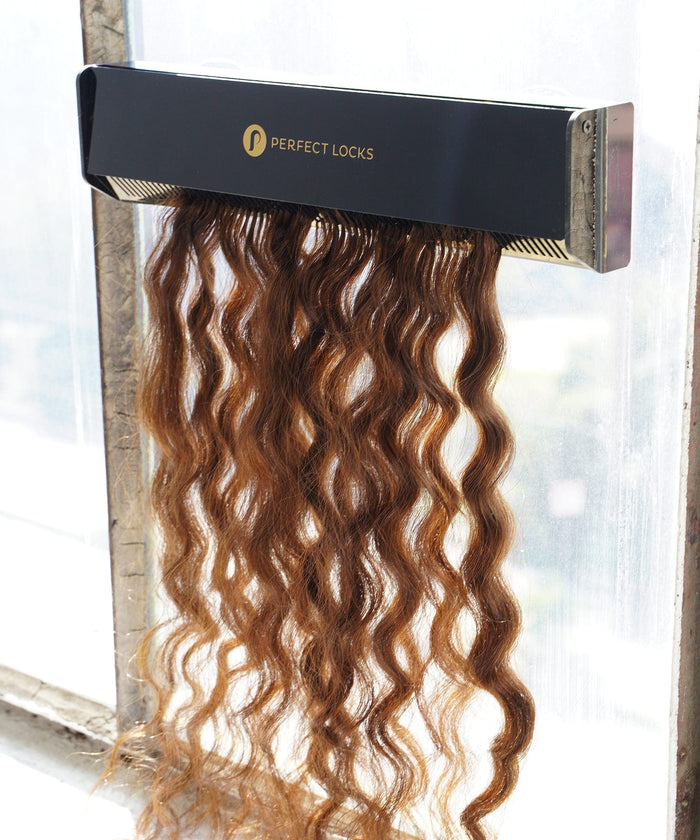 Wavy, brown hair extensions from Perfect Locks cascade from a sleek black box, illuminated by sunlight through a nearby window.