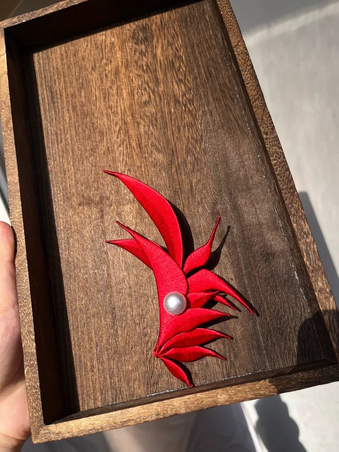 A red handmade Chanhua - style Zhang He hair accessory with a pearl in the center on a wooden box
