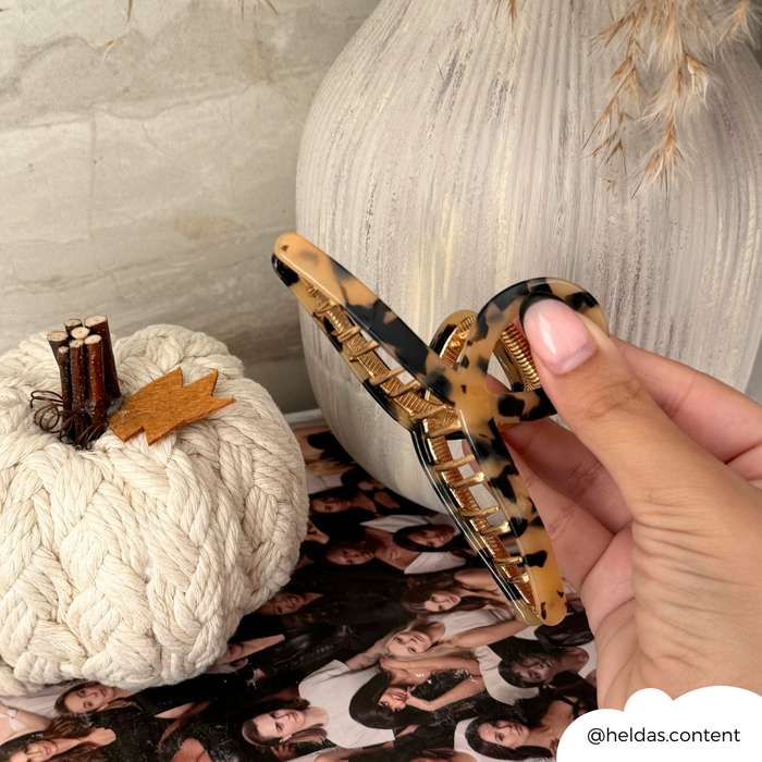 Hand holding an ivory tortoiseshell claw clip next to a small pumpkin and a white textured vase.
