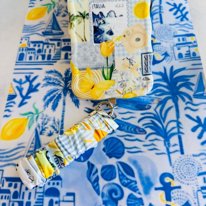 Blue and yellow La Dolce Summer scrunchie phone wrist strap in gingham and lemon print, clipped to the matching Amalfi Coast Wavy Luxe iPhone case with Italia and Capri travel stamps, styled on a blue-and-yellow Mediterranean palm and seashell print background.