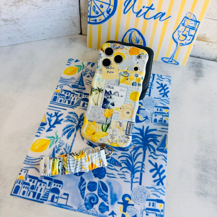 Blue and yellow La Dolce Summer scrunchie phone wrist strap in gingham and lemon print, clipped to the matching Amalfi Coast Wavy Luxe iPhone case with Italia and Capri travel stamps, styled on a blue-and-yellow Mediterranean palm and seashell print background.