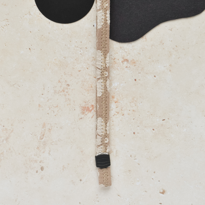 Mocha Flower Coconut Lane lanyard with a floral pattern on a speckled tan surface next to two abstract black shapes.