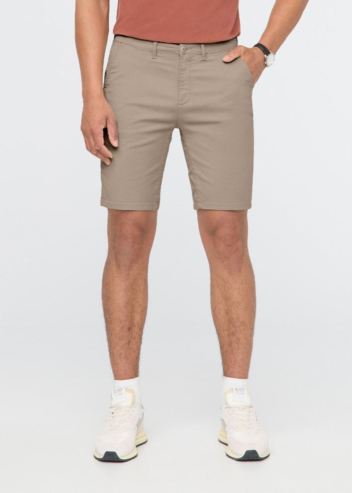 Sandstone-colored men's shorts with front pockets and flat front waistband, paired with rust-colored shirt and white sneakers