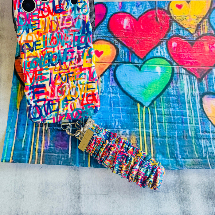 A vibrant Love Riot scrunchie phone wrist strap featuring a colorful neon graffiti "LOVE" and heart design attached to a matching Wavy Luxe phone case on a bright street art background.