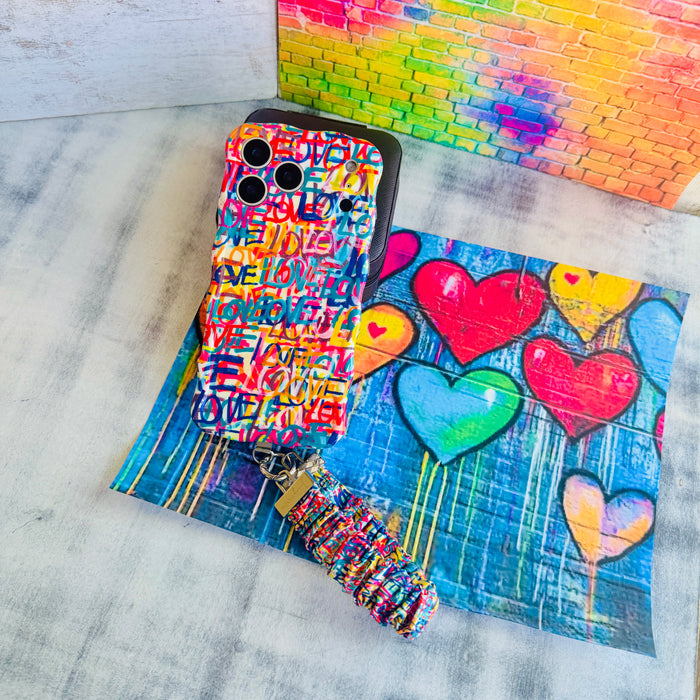 A vibrant Love Riot scrunchie phone wrist strap featuring a colorful neon graffiti "LOVE" and heart design attached to a matching Wavy Luxe phone case on a bright street art background.