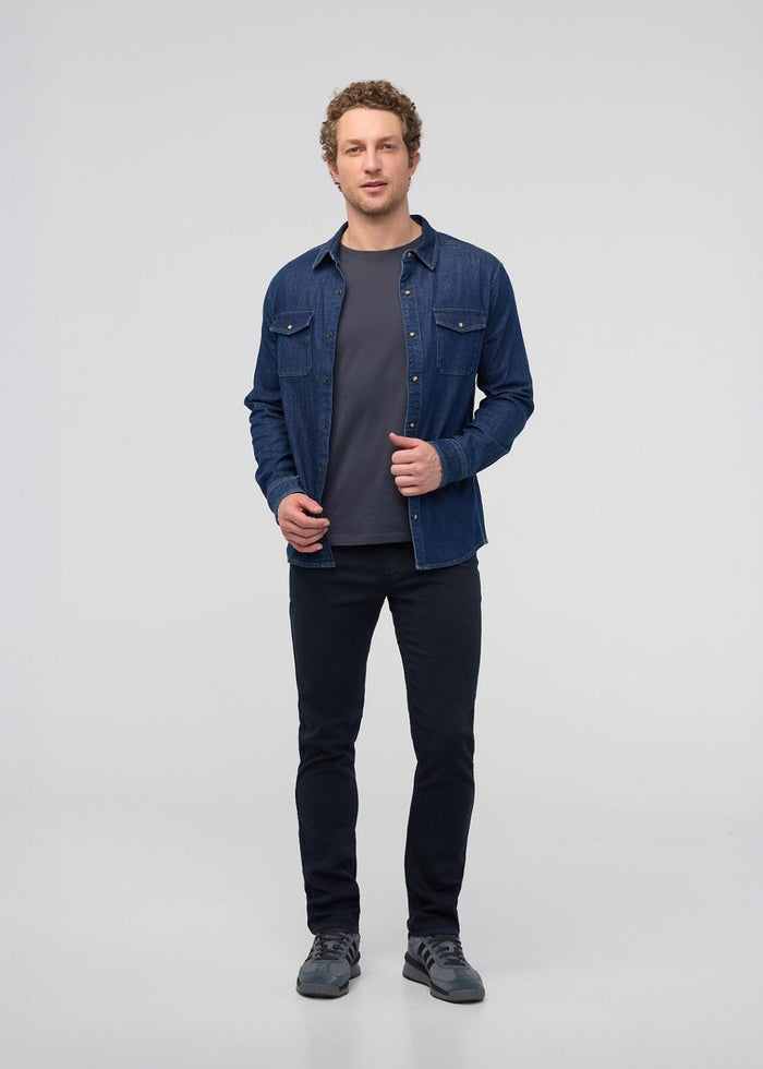 Man wearing a blue denim shirt over a gray t-shirt and black pants on a light gray background