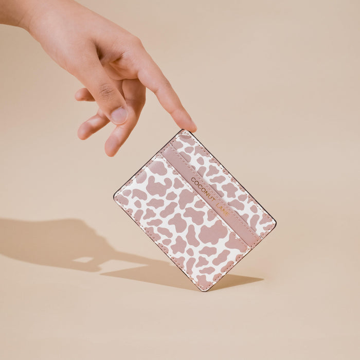 Hand holding a nude cow-print vegan leather card holder against a light beige background.