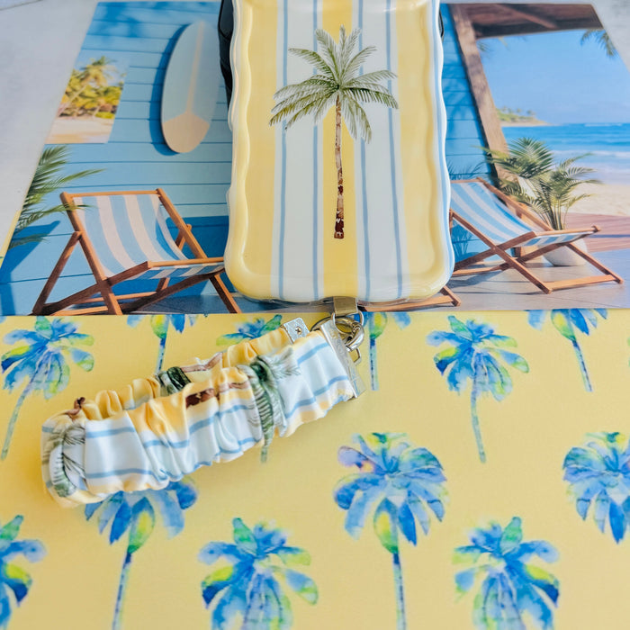 Yellow and blue striped Palm Shore Club scrunchie phone wrist strap with a palm-frond print, clipped to the matching coastal palm Wavy Luxe iPhone case, styled on a sunny yellow beach-club background with blue watercolor palm trees.