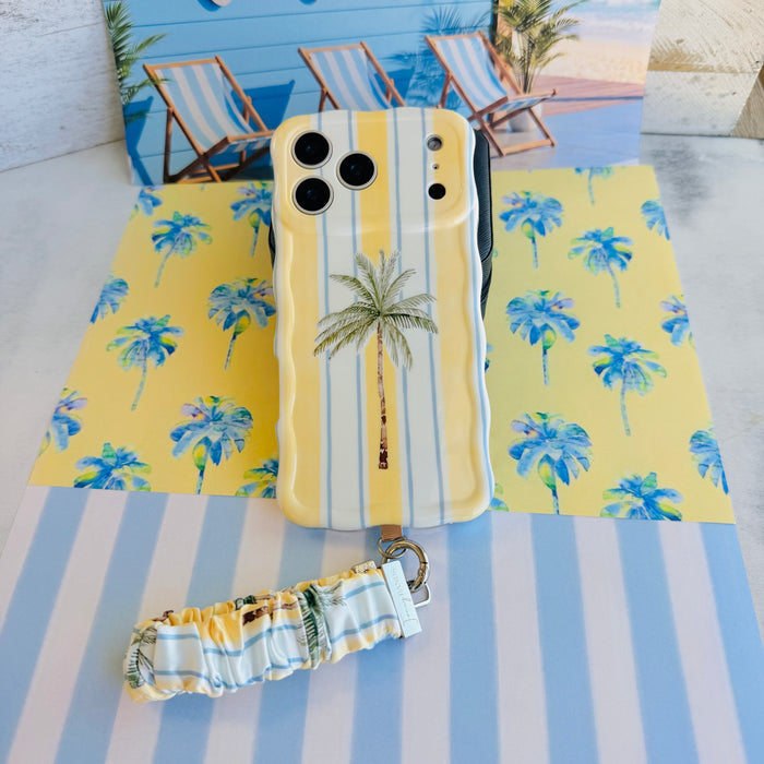 Yellow and blue striped Palm Shore Club scrunchie phone wrist strap with a palm-frond print, clipped to the matching coastal palm Wavy Luxe iPhone case, styled on a sunny yellow beach-club background with blue watercolor palm trees.