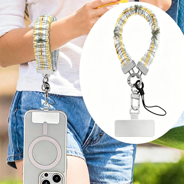 Palm Shore Club scrunchie phone wrist strap worn on the wrist with a phone in a clear MagSafe case, with an inset close-up of the yellow cabana stripe and palm print strap.