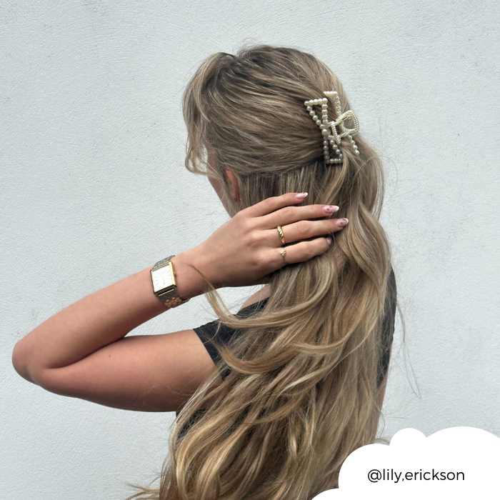 Woman with long wavy blonde hair wears a white pearl bow claw clip and gold jewelry against a plain white wall.
