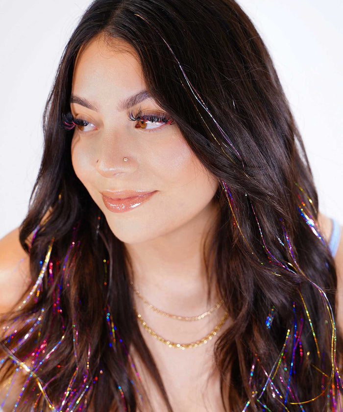 A woman with long dark hair smiles slightly, wearing rainbow hair tinsel, layered gold necklaces, glossy makeup, and a nose piercing.