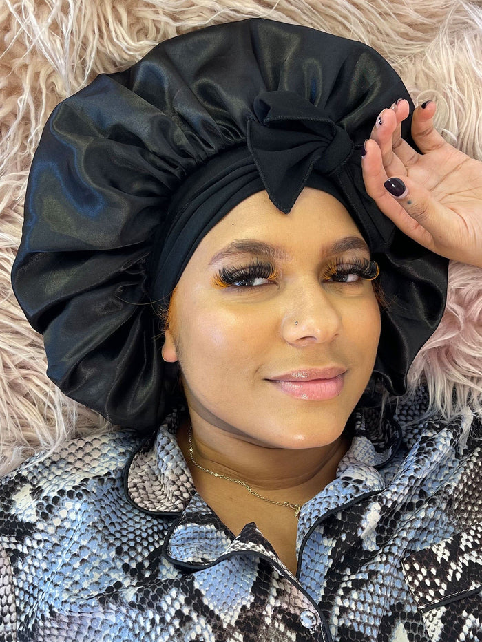 A woman relaxes on a fluffy surface, wearing a black satin bonnet and patterned pajama top, smiling softly in cozy nighttime comfort.