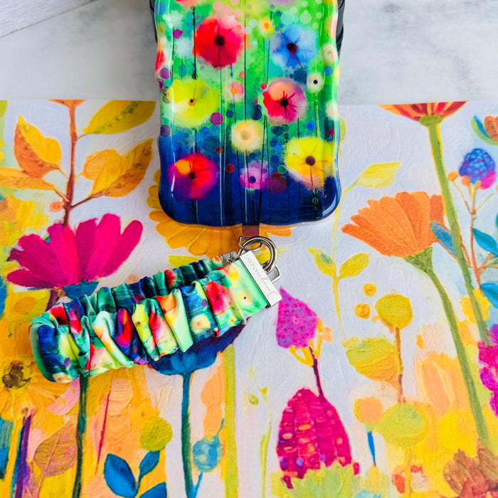 A vibrant Petal Princess scrunchie phone wrist strap in a colorful watercolor wildflower print, clipped to the matching Petal Princess Wavy Luxe iPhone case, styled on a bright painted floral background.
