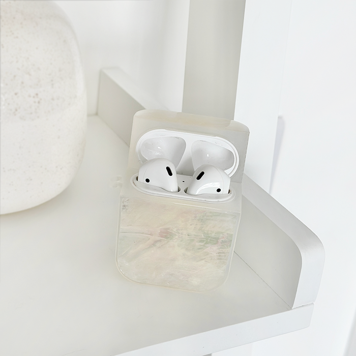 White real shell AirPods case with wireless earbuds on a white shelf next to minimal decor.