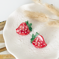 Pair of red Coconut Lane strawberry mini claw clips with white dots on a textured plate beside dried beige grass.