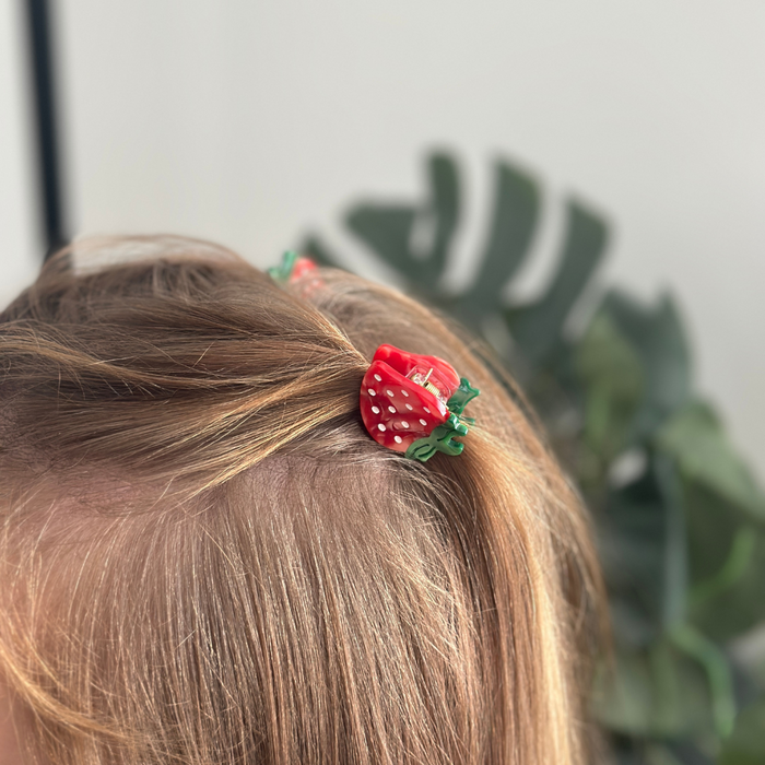 Child wearing Coconut Lane Strawberry Mini Claw Clips in hair, with a green leafy plant blurred in the background.