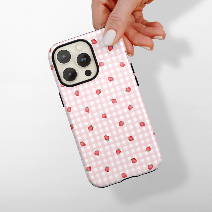 Hand holding Coconut Lane tough phone case with pink gingham pattern and small red strawberries.
