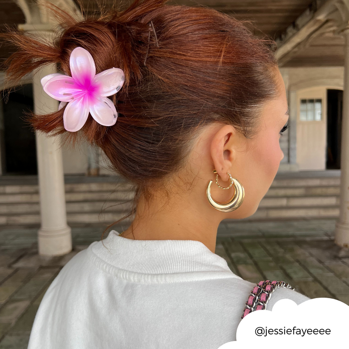 Woman with brown hair in a bun wearing a pink floral claw clip, gold hoop earrings, and a white shirt outdoors.