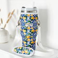 A vibrant Coconut Lane Tumbler Carry Case - Amalfi Lemon, adorned with a charming floral and fruit pattern featuring lemons and green leaves on a blue background. It includes a coordinated strap and an attached small pouch. In the background, there is a decorative vase filled with dried wheat stalks resting on a white surface.