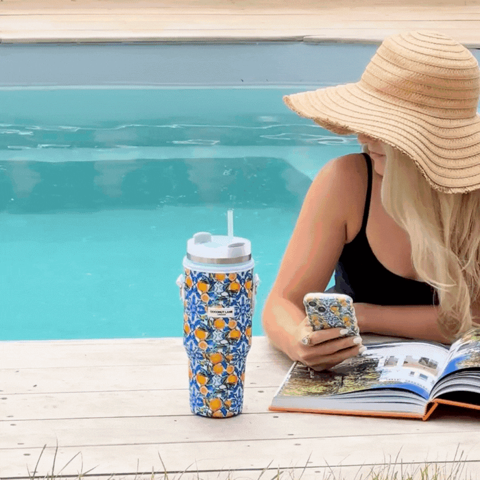 Woman in a sunhat reads by a pool with a yellow Coconut Lane tumbler carry case featuring an Amalfi lemon pattern nearby.