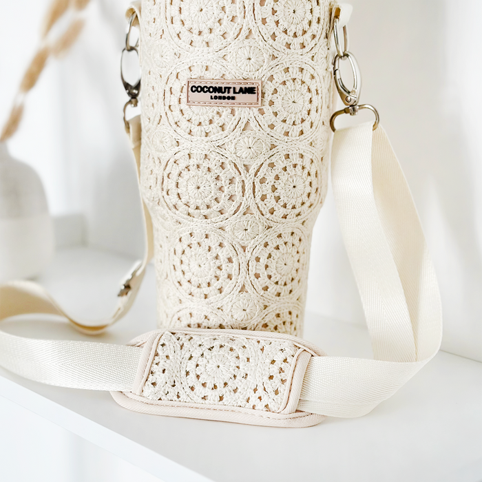 The Tumbler Carry Case - Festival Crochet by Coconut Lane is displayed. This beige crochet water bottle holder showcases intricate circular patterns and includes a cream-colored adjustable strap with a padded section for added comfort. The travel accessory features a branded label reading "COCONUT LANE LONDON." It is placed on a white surface.