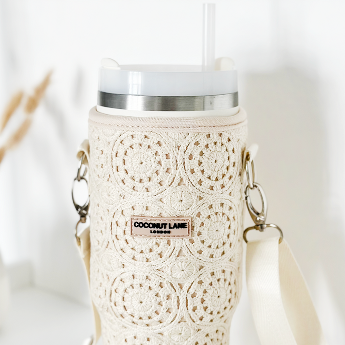 A close-up of a white tumbler protected by the Tumbler Carry Case - Festival Crochet by Coconut Lane, featuring a textured, intricately crocheted sleeve with floral patterns. The tumbler has a white lid with a transparent straw and beige shoulder straps attached on both sides, making it the perfect travel accessory. A label on the sleeve reads "Coconut Lane London.
