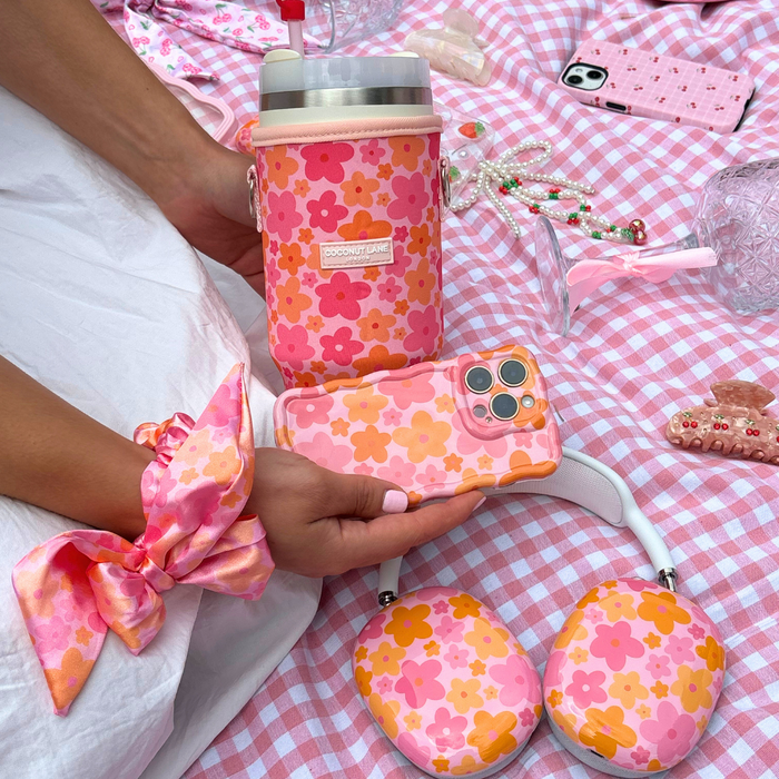 A person is holding an iPhone with a pink and orange floral case, perfectly matching their Coconut Lane Tumbler Carry Case - Pink & Orange Bloom. Also seen is a hair scrunchie, headphones, and various accessories laid out on a pink and white checkered picnic blanket.